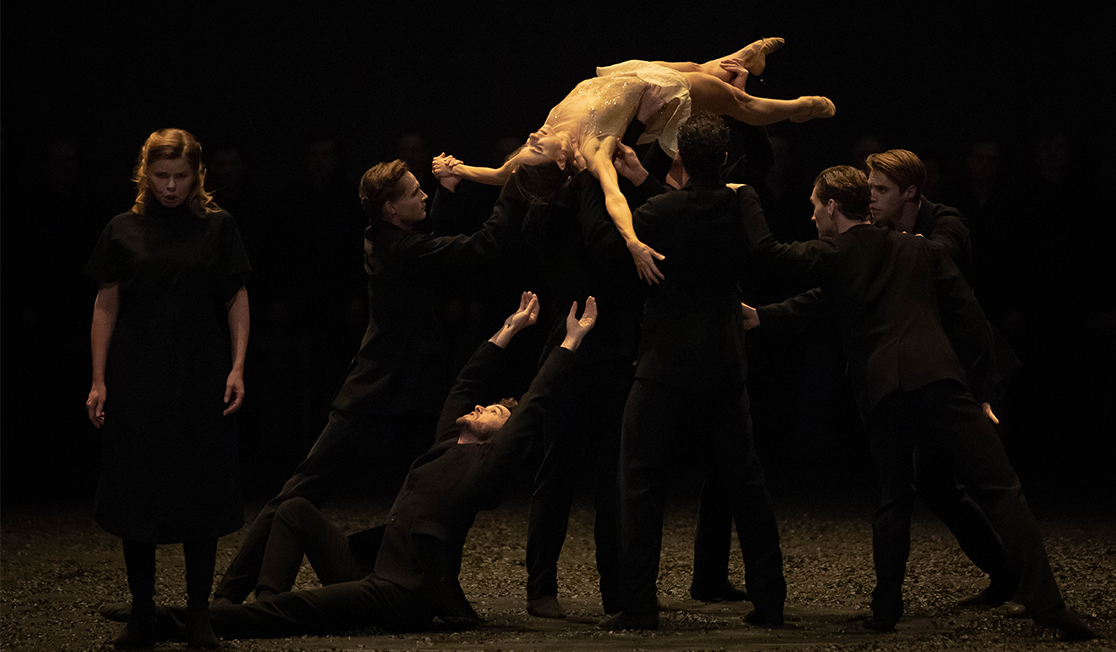 Dancers wearing black lift a dancer wearing a sheer slip dress into the air. The background is dark and beside them is a person singing 