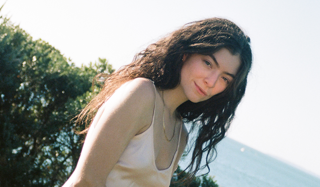 A woman with long brown wavy hair looks at the camera with a half-smile. She is wearing a gold chain and a slip dress and there is foliage and water visible behind her. The photo is angled to the side rather than straight on.