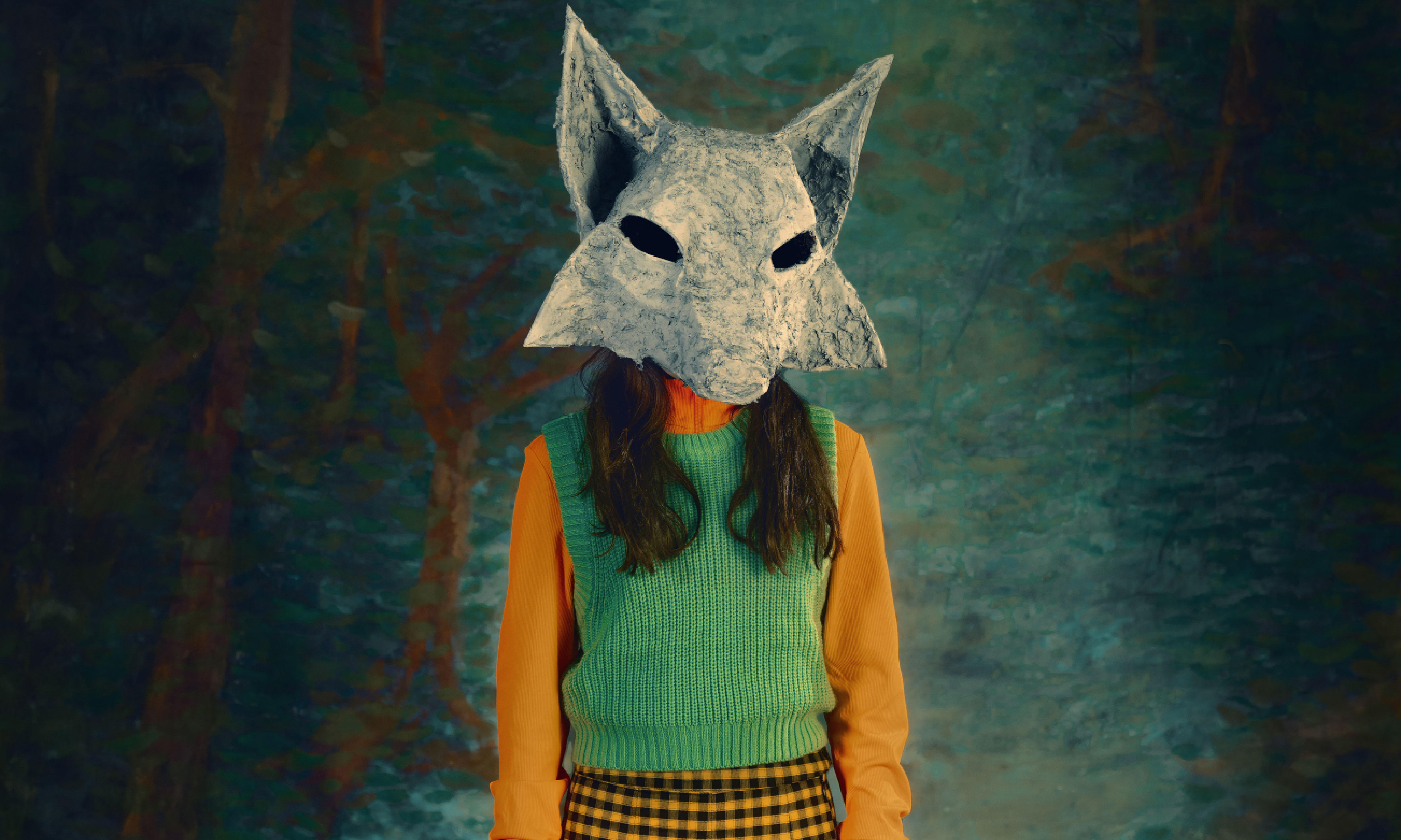 A child stands in front of a forest wallpaper, they are wearing a simple wolf mask with dark eyes and a vibrant green vest, yellow skirt and high socks. 