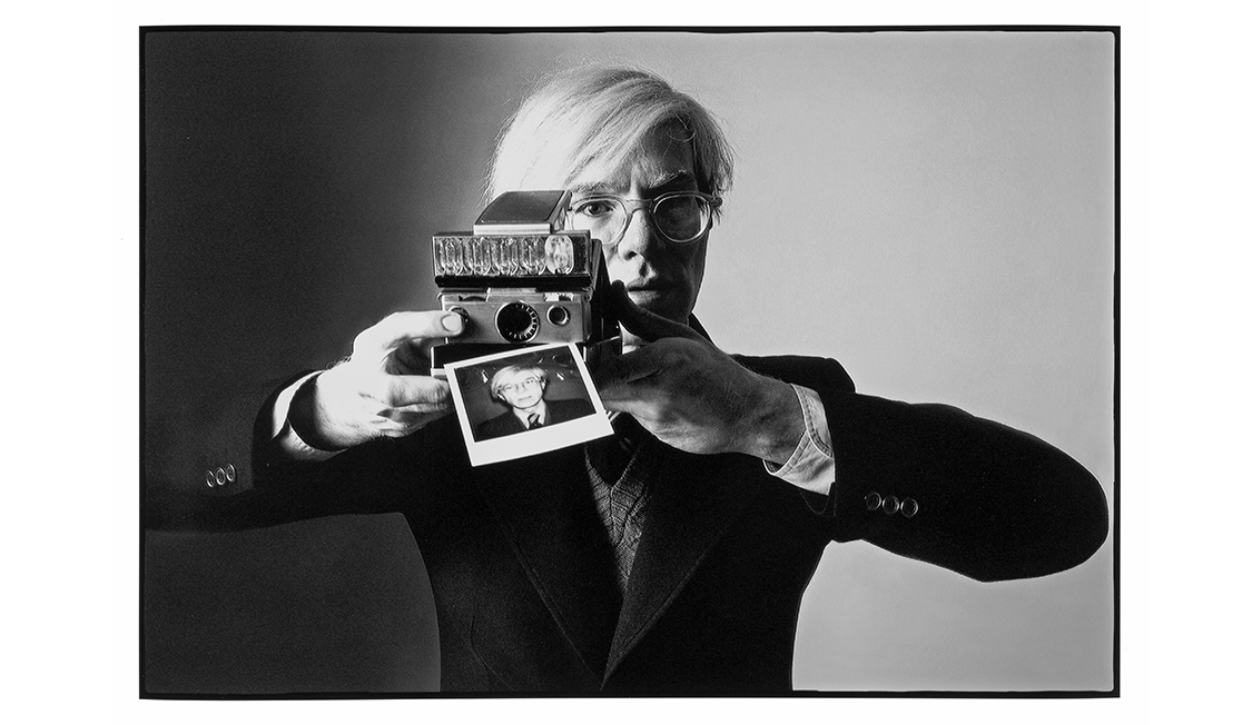 A black and white image shows a man with white hair and glasses wearing a suit and holding a polaroid camera with an image of himself being printed from it.