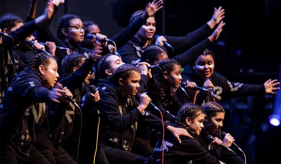 A group of young people hold microphones and are dressed in black, they have braids in their hair and have their arms in front of them