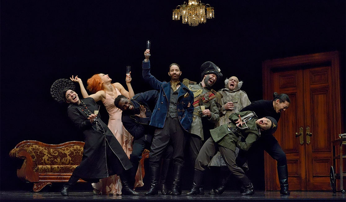 A group of dancers wearing formal military attire are making exaggerated laughing expressions and the person in the centre of the group is pointing a pistol into the air. 