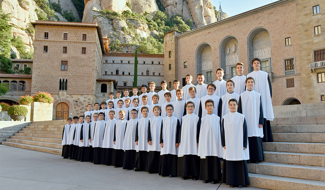A group of about 30 young boys stand, smiling on the stone steps of the Montserrat monastery. They are wearing black and white gowns. Behind the monastery is a large rocky mountain.