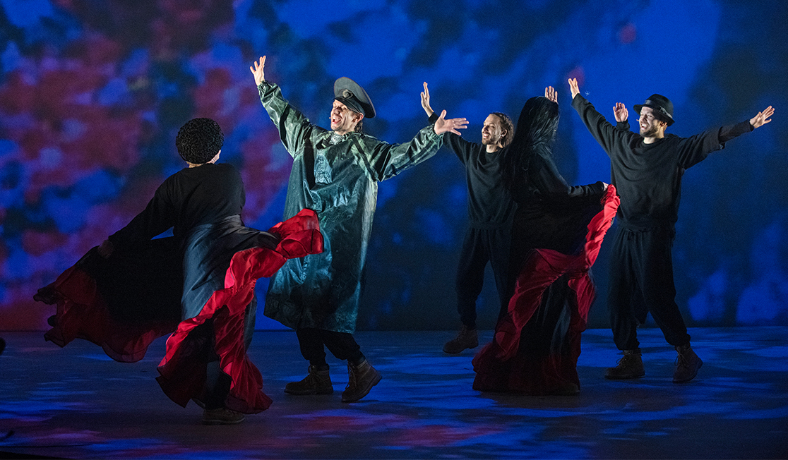 Performers are shown on stage with the blue background, two people are wearing black and red skirts that are billowing, three people have their arms up and are smiling. The man at the front wears a long jacket, military hat and boots. 