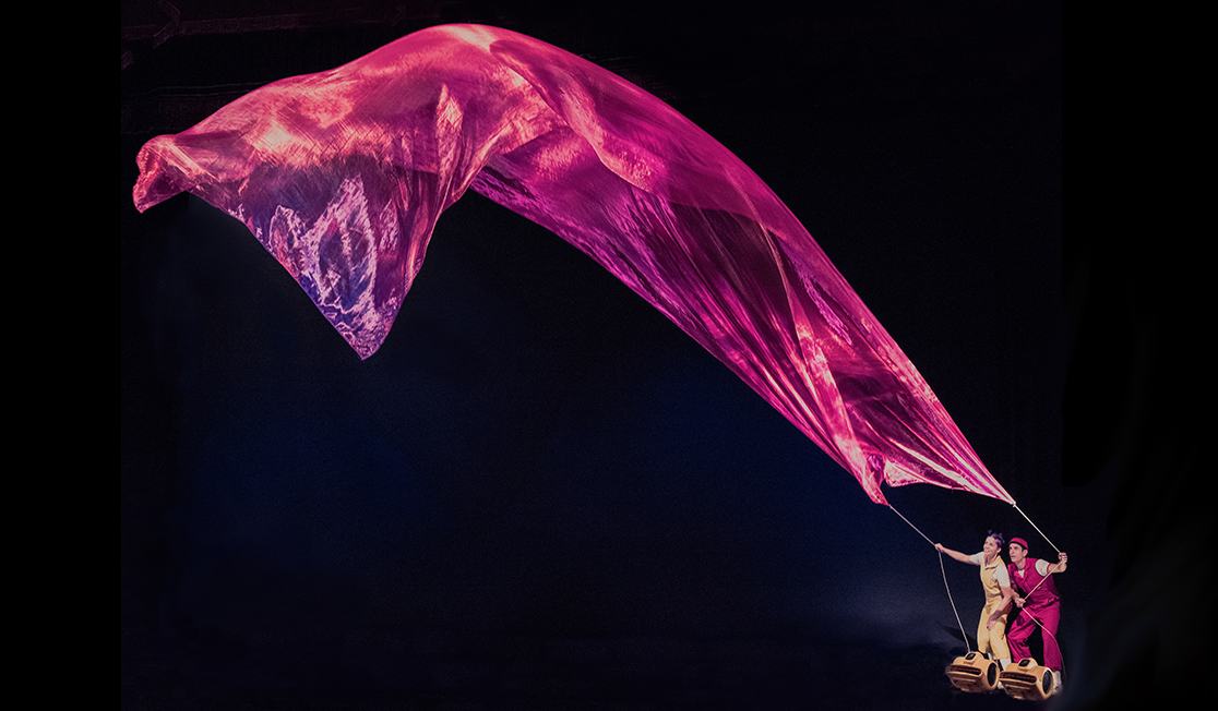 Two people on a dark stage holding a huge piece of iridescent fabric like a kite. It flies above them. One person wears a yellow jumpsuit and has purple hair, the other wears a red jumpsuit and has a red headband.