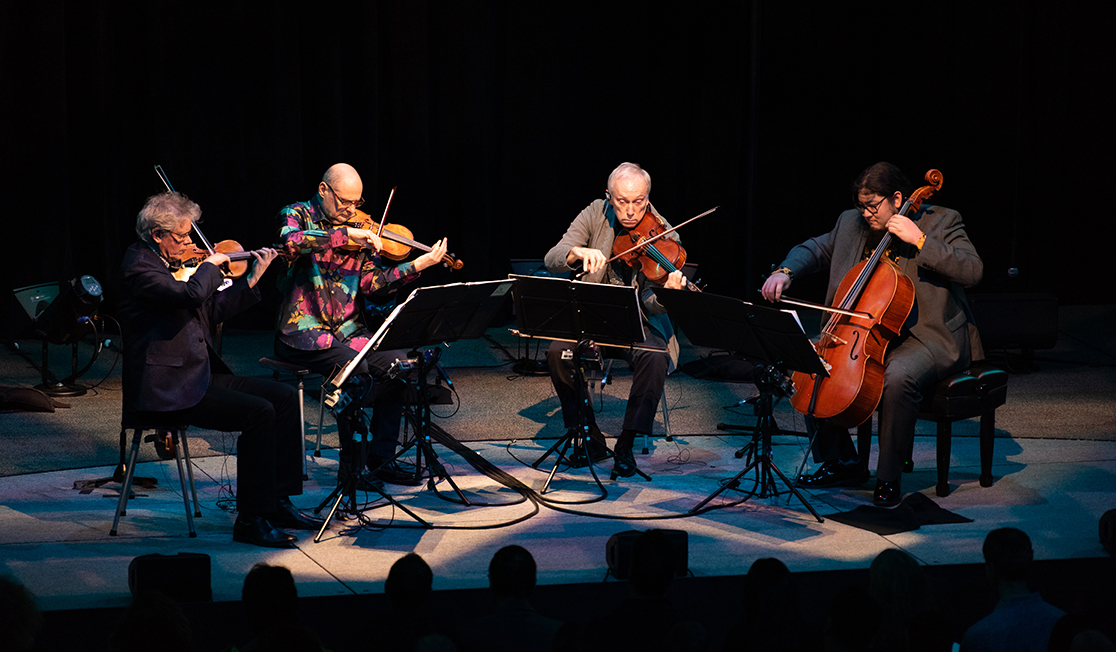 Four men sit on a darkened stage playing string instruments.
