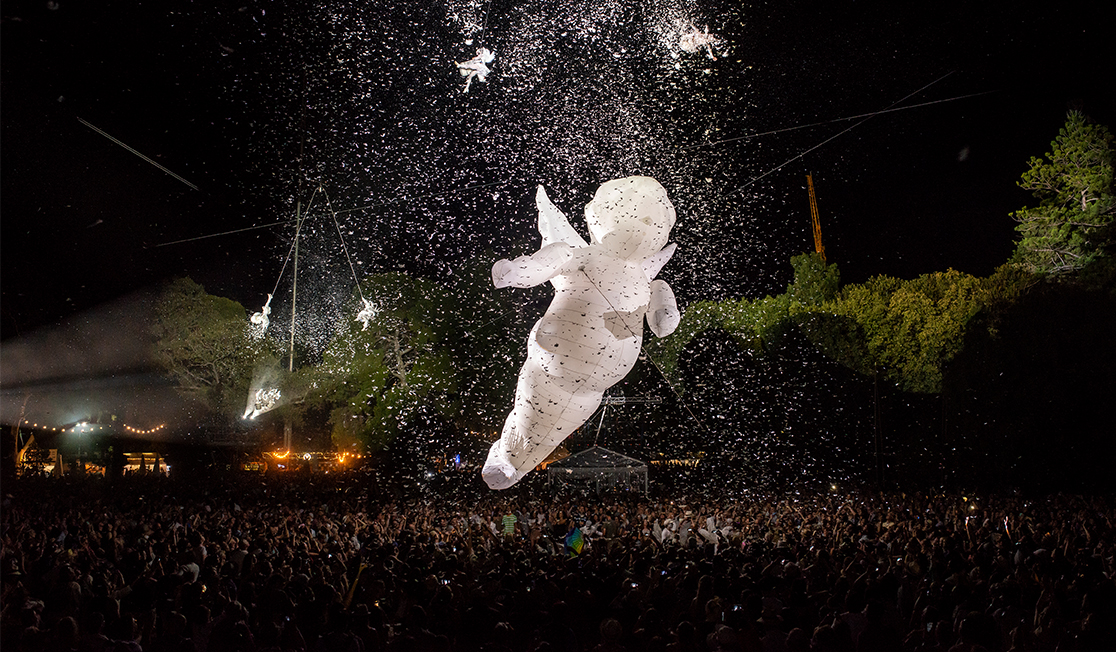 A giant balloon shaped like an angel suspended in the air over a crowd at night time.