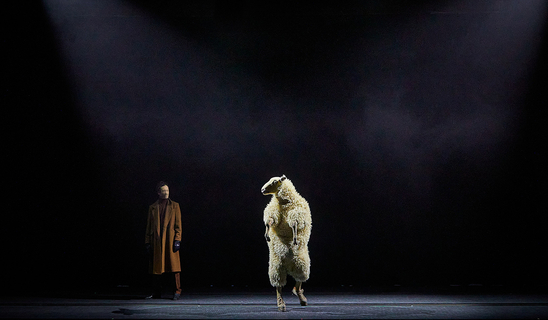A man in a brown coat and a person dressed in a sheep costume stand on a darkened stage.