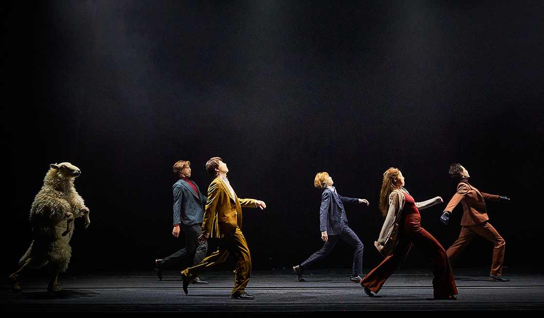 A person in a sheep costume walks behind a group of people dressed in bright colours.