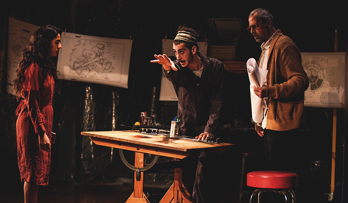 Three people stand in a dark room around a wooden desk, paper maps are pinned on a string around them. A man stands at the desk and points into the distance. A man in a brown jacket and a woman in a red dress are watching him.