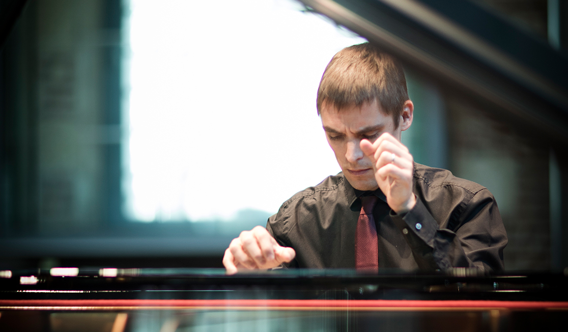 Cédric wears a black shirt and a red tie, he has short brown hair. He is sitting at a piano and his hands are raised as he plays. He has a concentrated expression.