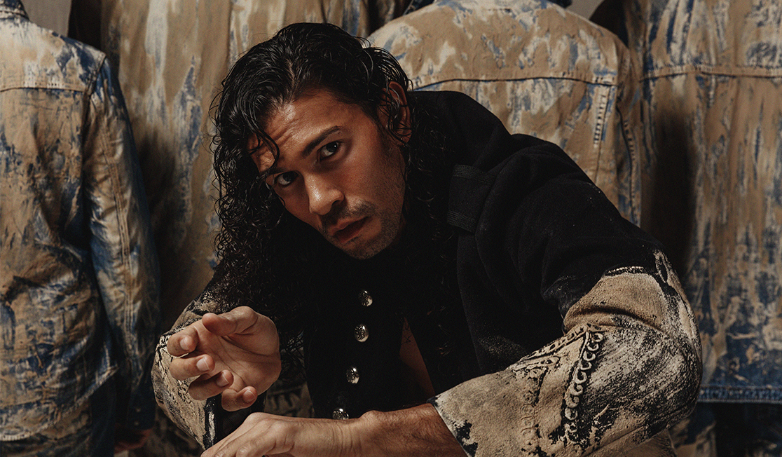 A man in a black jacket with mud on the sleeves leans towards the camera, he has long curly, dark hair and a serious expression, there are people standing behind him and facing away, they are wearing denim jackets covered in drying mud.