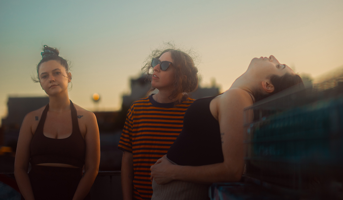 Three people stand outdoors in an industrial area, the person on the left wears a tank top and has dark hair in a bun, the person in the middle wears a striped orange t-shirt and sunglasses and the person on the right wears a black tank top and leans backwards over a concrete structure.