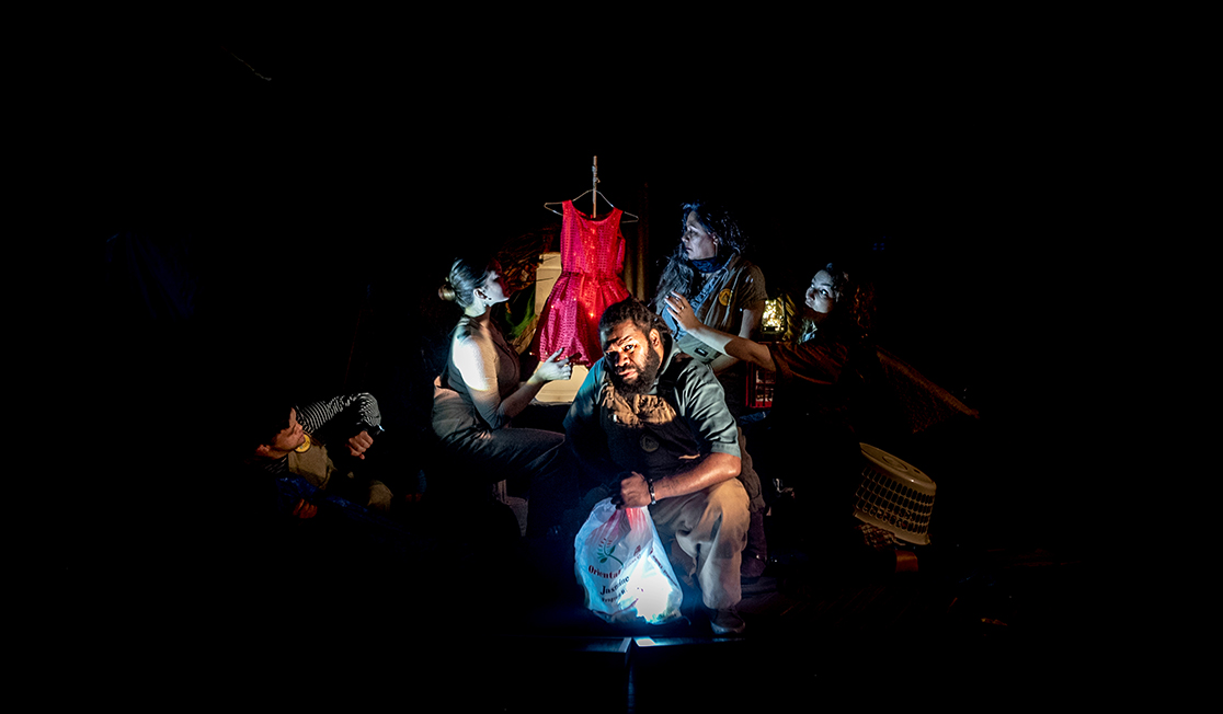 A group of 5 people sit in a very dark room, the man at the front holds a plastic bag and looks straight ahead, the other people are looking towards a small red dress on a coat hanger. Around them is a washing basket and a plastic crate.