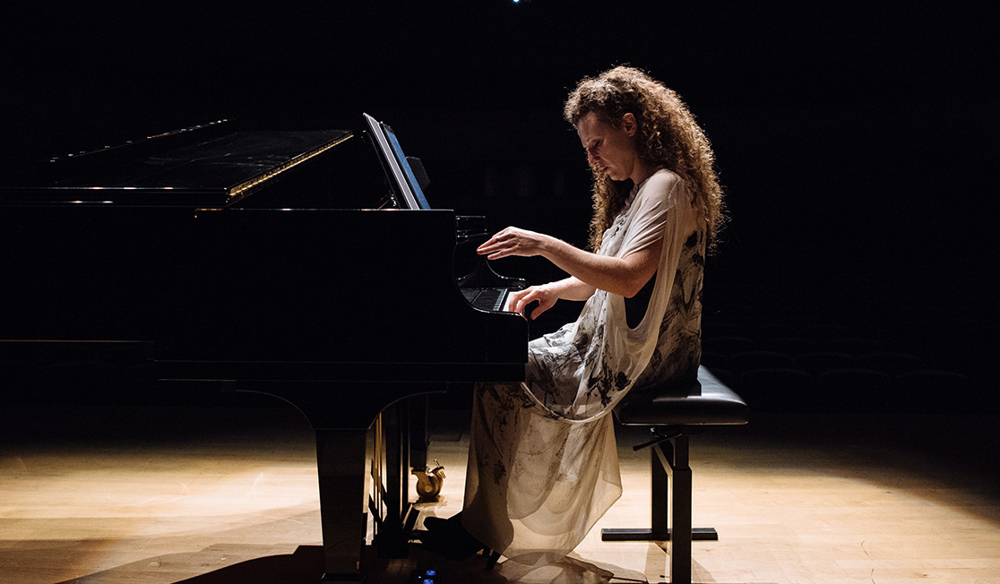 A woman with brown curly hair sits at a piano on a stage, she is wearing a long sheer dress and is looking at the keys as she lifts one hand. 