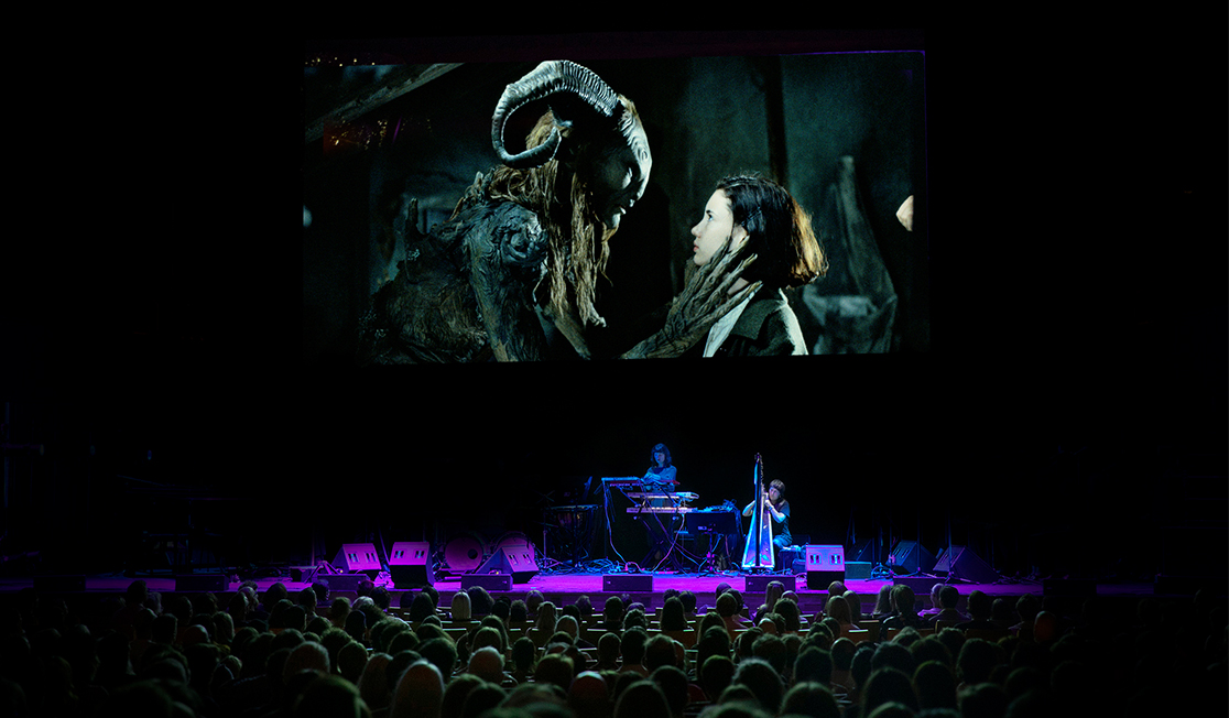 A crowd sits in a dark theatre facing the stage. On stage there is a musician standing behind a mixing desk and a screen behind them showing a scene from a film with a creature with curled horns speaking to a young girl.