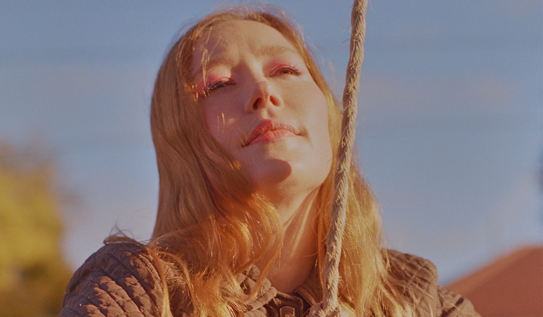 A woman with red hair is pictured in a brown jacket and is wearing pink eyeshadow. She is tilting her face up and holds a rope in front of her connected to a tyre swing.