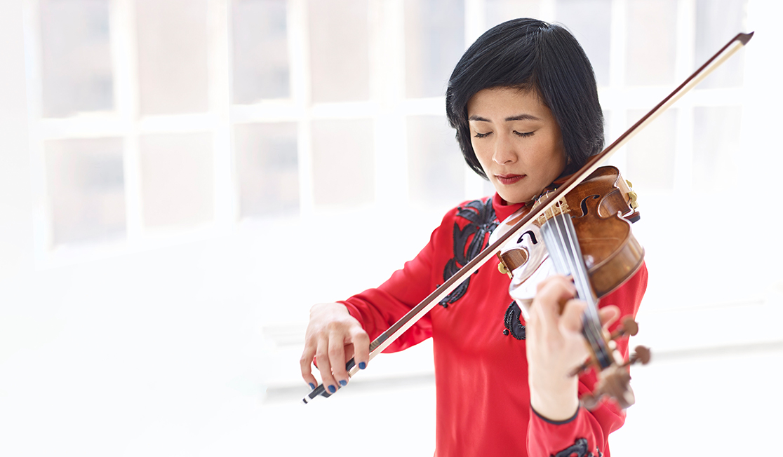 A woman with dark hair is shown standing and playing the violin in front of a bright white background. She wears a red blouse with black embellishments and she has her eyes closed.