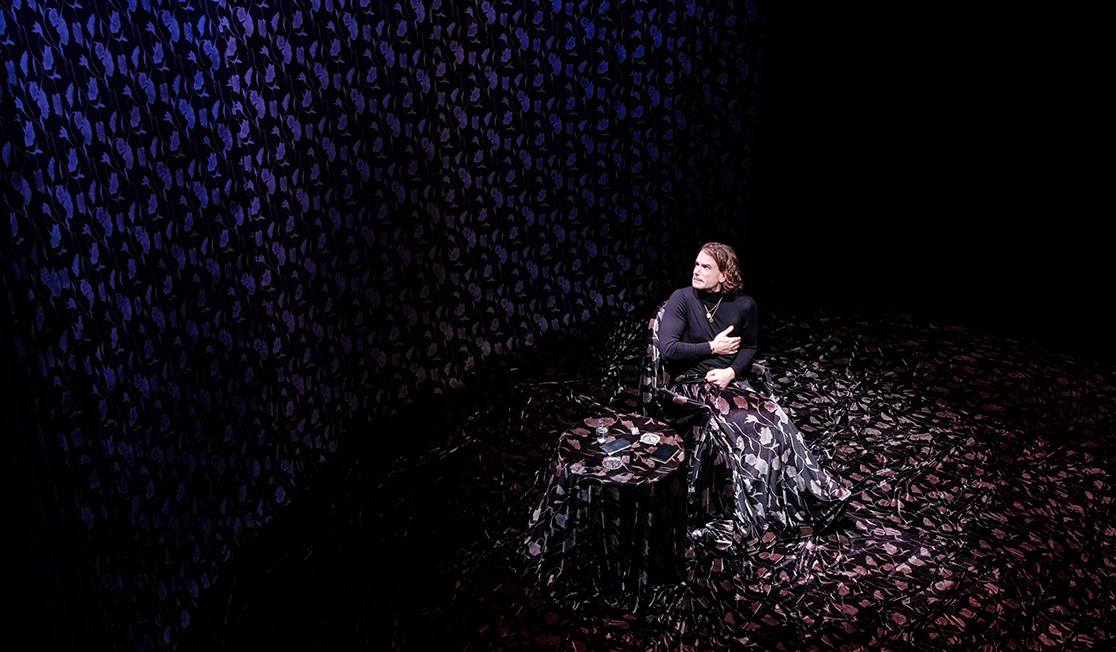 An actor sits in a dark room wearing a floral silk skirt that goes beyond their feet and covers a table and goes up the walls. They have a worried expression,