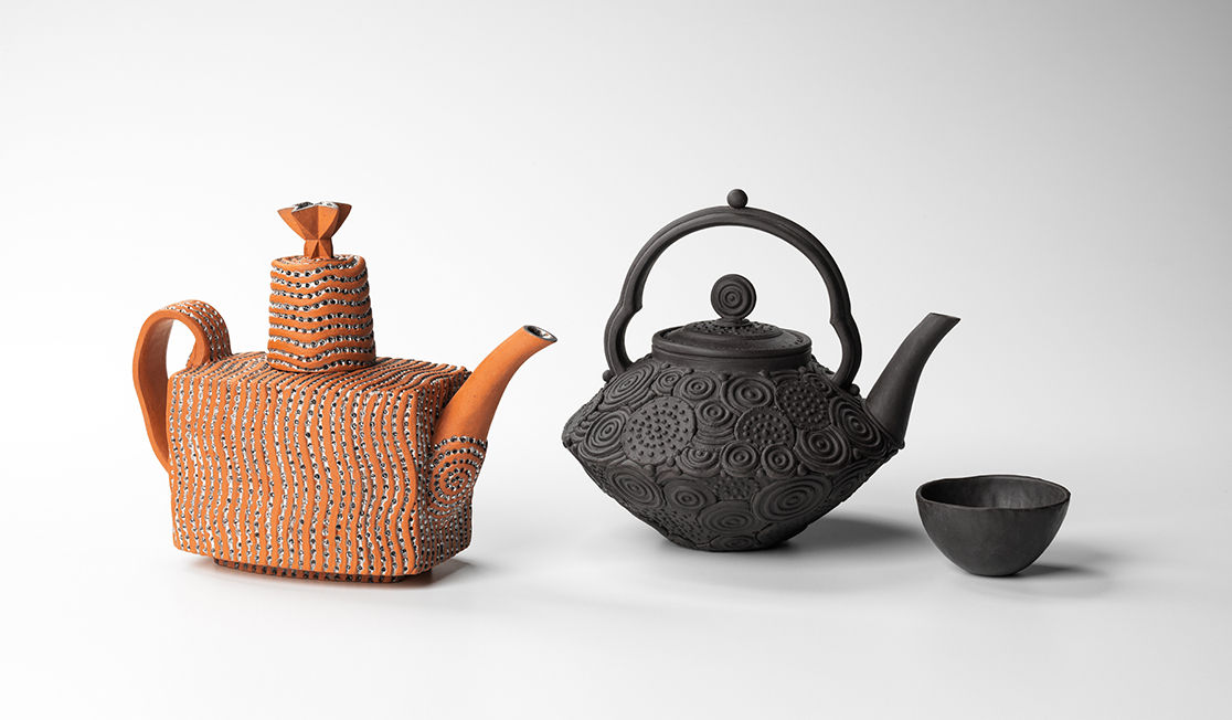 Two teapots are in front of a white background. One is orange and has spots and is more of a rectangular prism shape. The other is black and has swirls and is more circular. It has a small smooth black tea bowl in front of it.