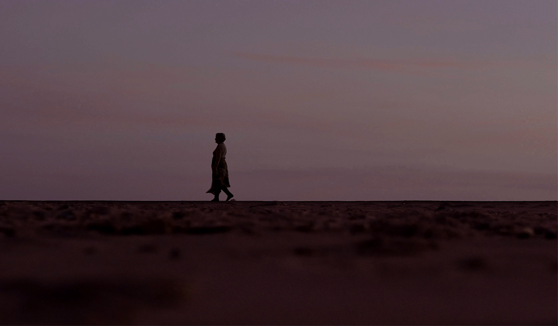 A person is walking along a flat landscape with a dark pink sky behind them.