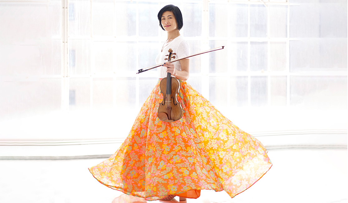 A woman in a bright white room holds a violin and wears a billowing orange skirt and a white top. She has dark hair and is smiling.