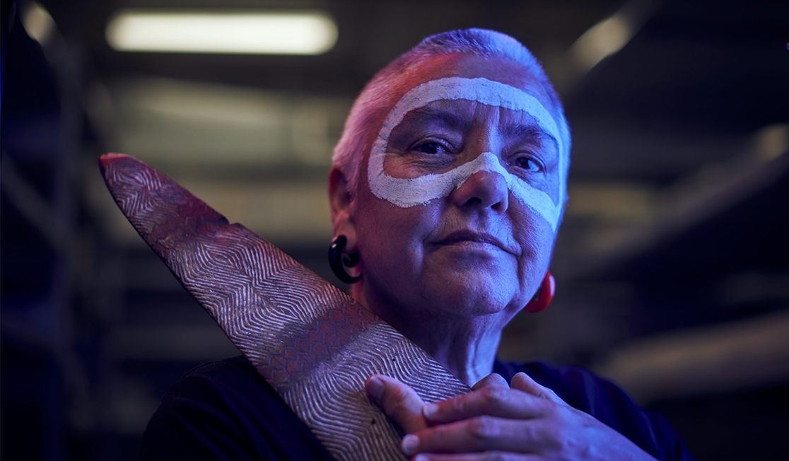 A woman illuminated in blue light faces ahead. She has a buzz cut and white paint around her eyes. She holds an intricately carved piece of wood and has a gentle expression.