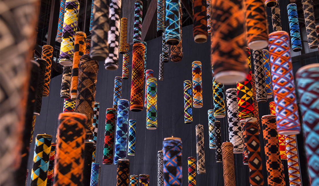 Brightly coloured and patterned cylinders hang from clear wire on a dark grey background. They appear to be small lanterns or rolls of fabric.