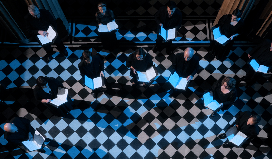 A bird's eye view shows a choir of 13 people wearing black and holding music scores in front of them. They are standing on a checkerboard patterned floor and it is night time.