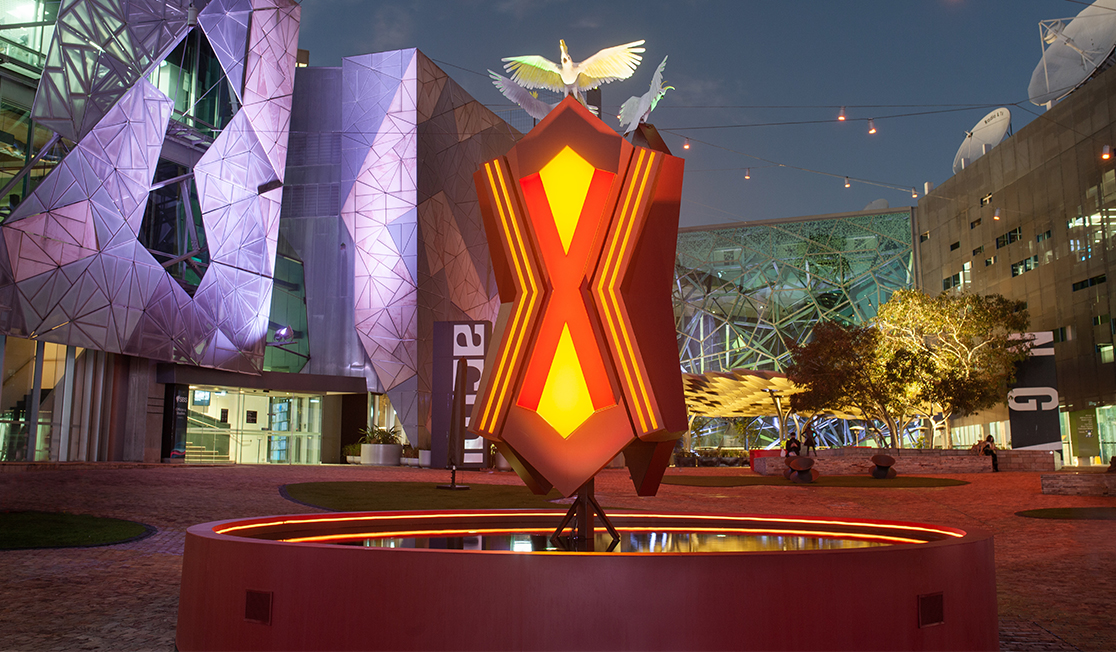 A large glowing structure is shown in a dark area surrounded by buildings. A red and yellow geometric shape is on a round stand. It has sulphur crested cockatoos with open wings on the top.