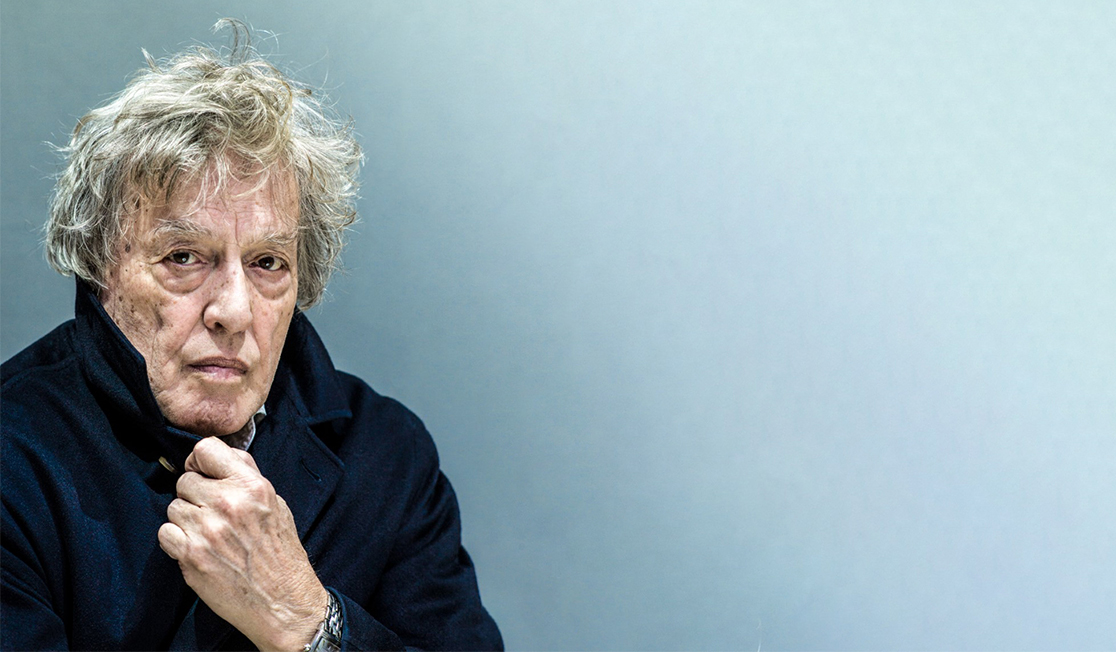 A man with grey hair wears a blue coat and holds the collar with one hand. He has a serious expression and is in front of a blue background.