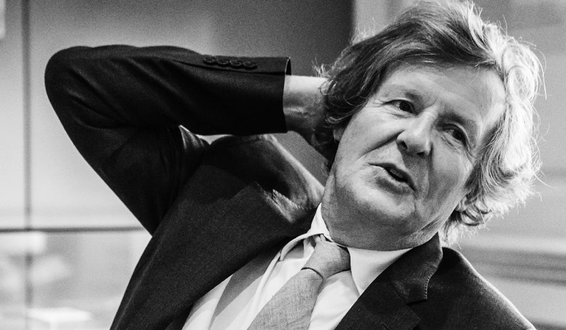 A man in a black and white photo is wearing a suit and looks to be in conversation. He has his hand on the back o f his head and is looking off to the right.