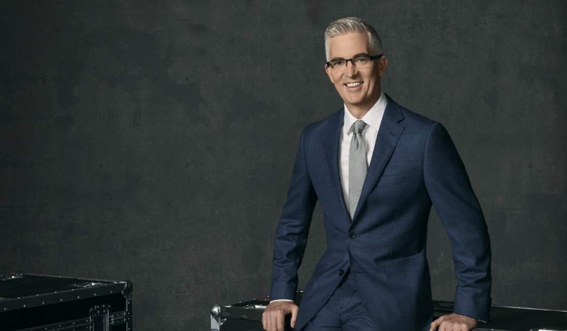 A man in a blue suit and a grey tie sits on a chrome and black storage box. He has grey hair and glasses and is smiling.