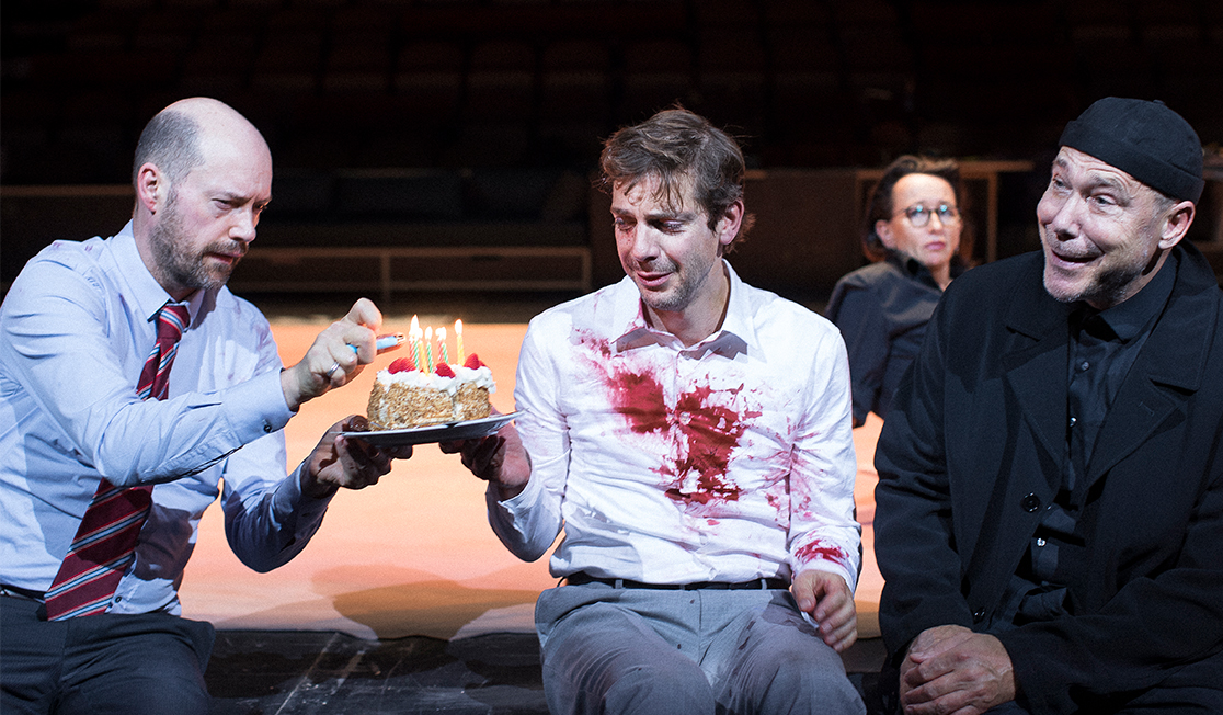 Three men sit on stage. One wears a shirt and tie and is lighting candles on a birthday cake, the man in the middle wears a white shirt with blood stains on the chest, the man on the end wears all black and is smiling.