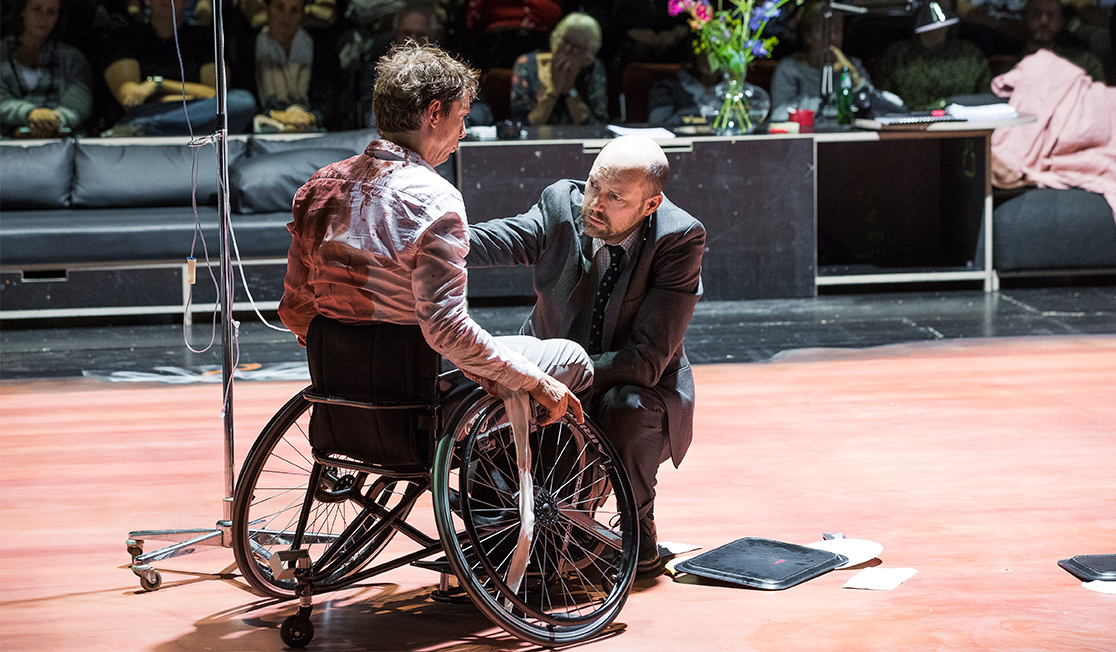 On stage, a man is in a wheelchair with an IV drip, he is wearing a shirt that is stained with blood. A man in a suit has his arm on the shoulder of the man in the wheelchair.