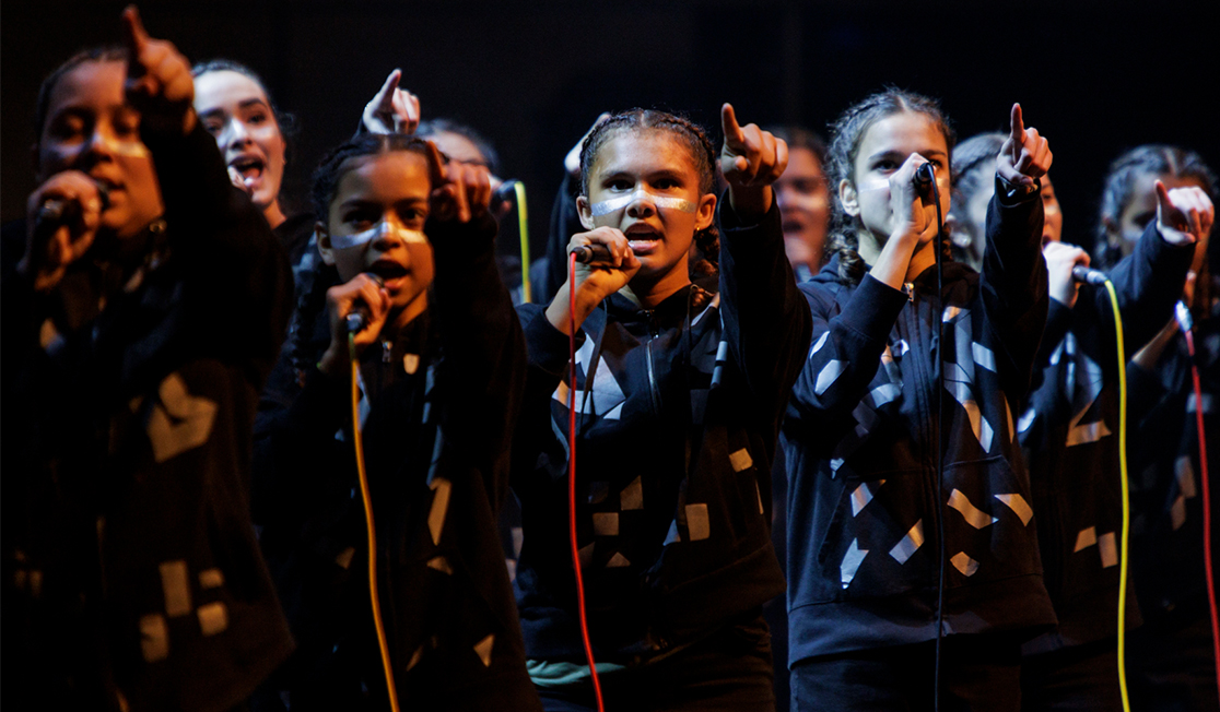 A group of young people hold microphones and are dressed in black, they have braids in their hair and are pointing in front of them