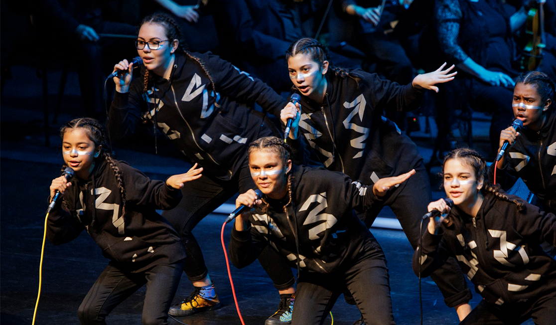 A group of young people hold microphones and are dressed in black, they have braids in their hair and lean to the left as they dance 