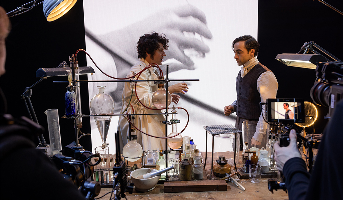 Two men stand behind a table covered with lab equipment, it has test tubes, a bunsen burner and bottles and vials. Behind them is a screen with an image of a hand. 