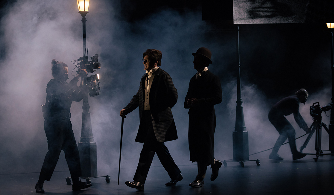 Two men in formalwear walk on a dark stage lit by street lamps. In front of them and behind them are crew members wearing black and holding cameras and lights.
