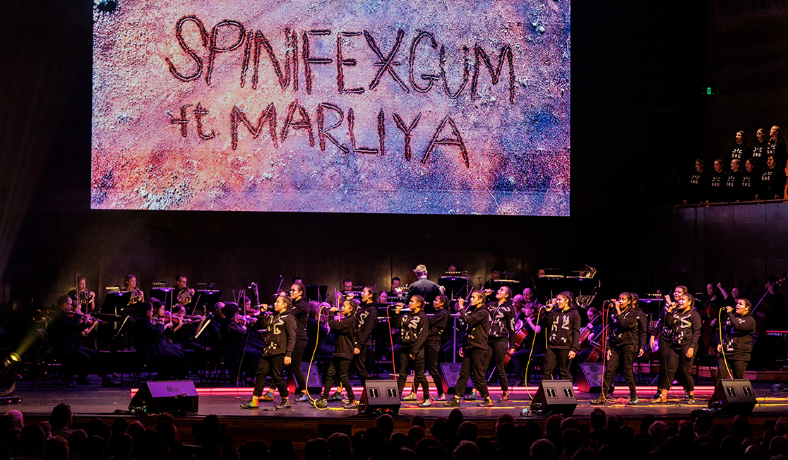 A group of young people sing on stage with an orchestra behind them and a screen overhead that reads 'Spinifex Gum ft Marliya'