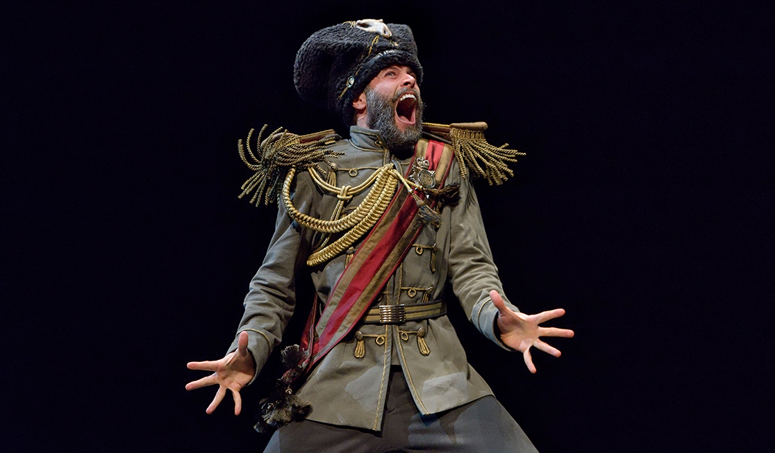 A man in a military style costume screams with his hands splayed on front of him in front of a black background.