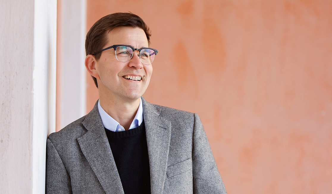 Paavali Jumppanen, a white man with dark brown hair and glasses, is wearing a grey blazer and smiling while looking to the right of the camera in front of a neutral terracotta-coloured background.