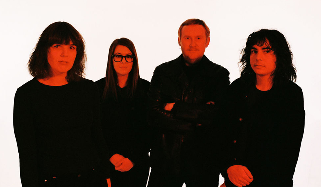 Four people wearing black look at the camera with serious expressions, bathed in a reddish light.