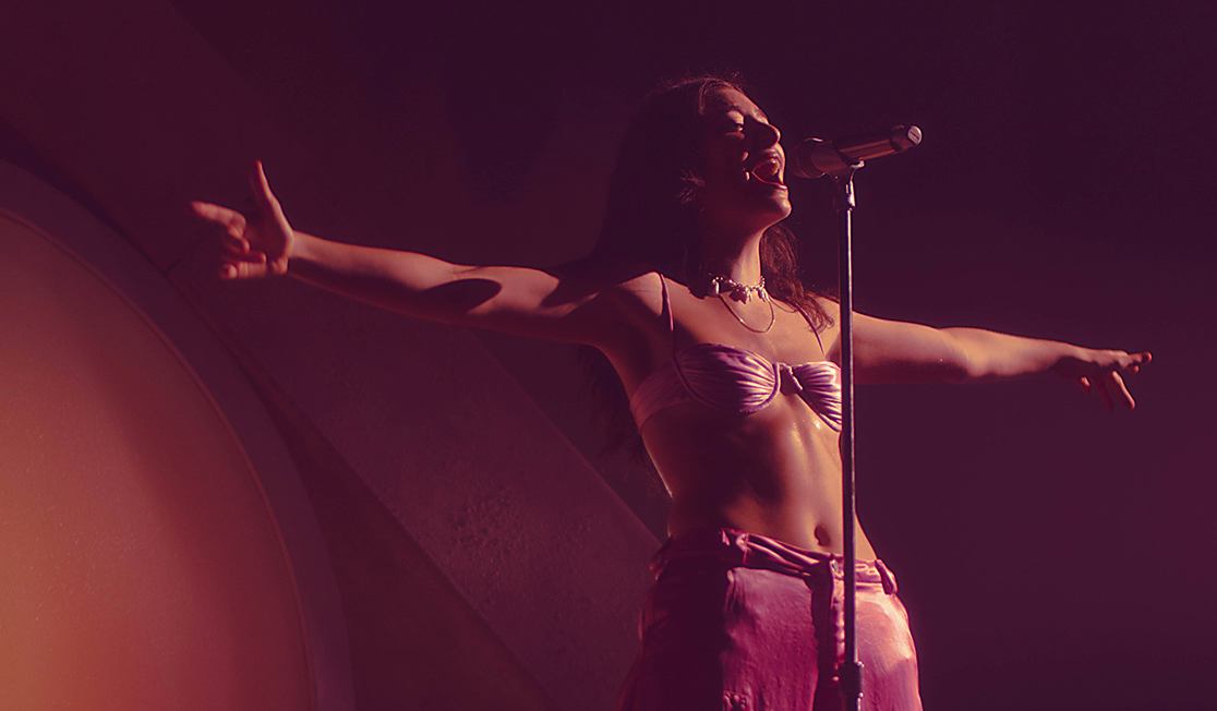 A woman with dark brown hair wearing a pink bikini top and pink pants stands with arms outstretched while singing into a microphone.