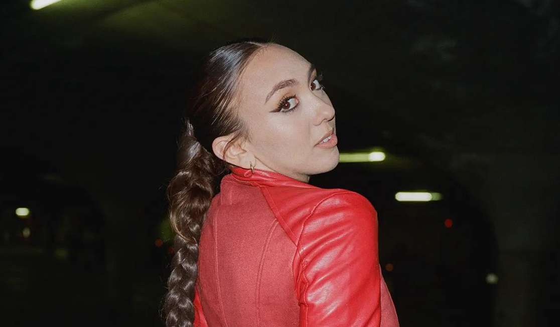 A young woman with a long brown braid and wearing a red jacket looks over her shoulder at the camera with a slightly surprised expression.