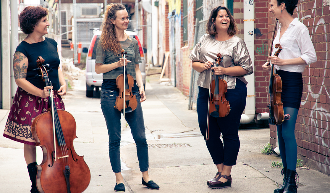 Four women stand in an alley while holding string instruments. They are looking at one another and smiling.