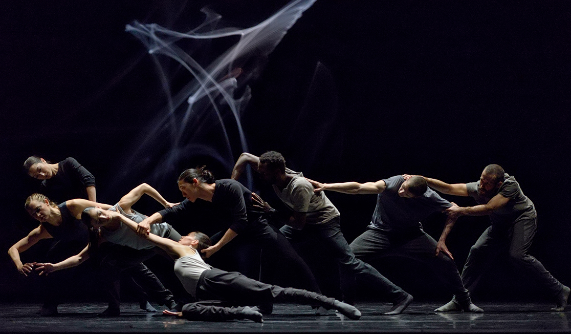 Dancers dressed in grey and black move together through dance movements with serious expressions.