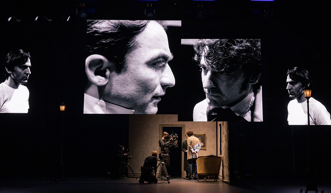 Screens showing black and white footage of two men from different angles are displayed above live action on a stage.