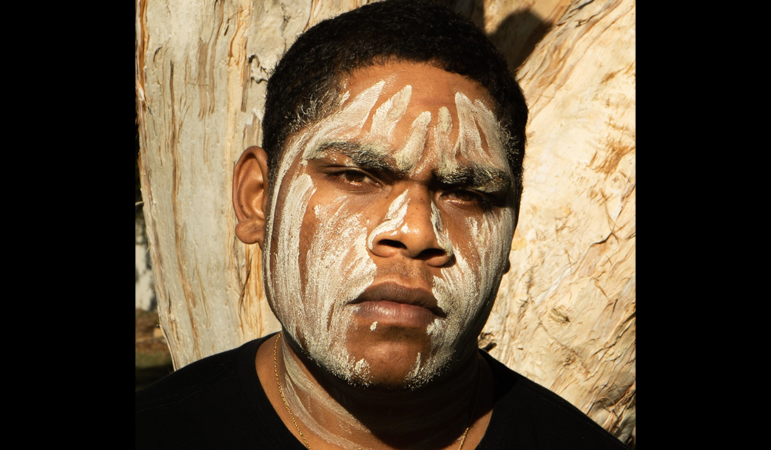 A young man with short black hair looks at the camera with a serious expression. He is wearing a black t-shirt and white face paint.