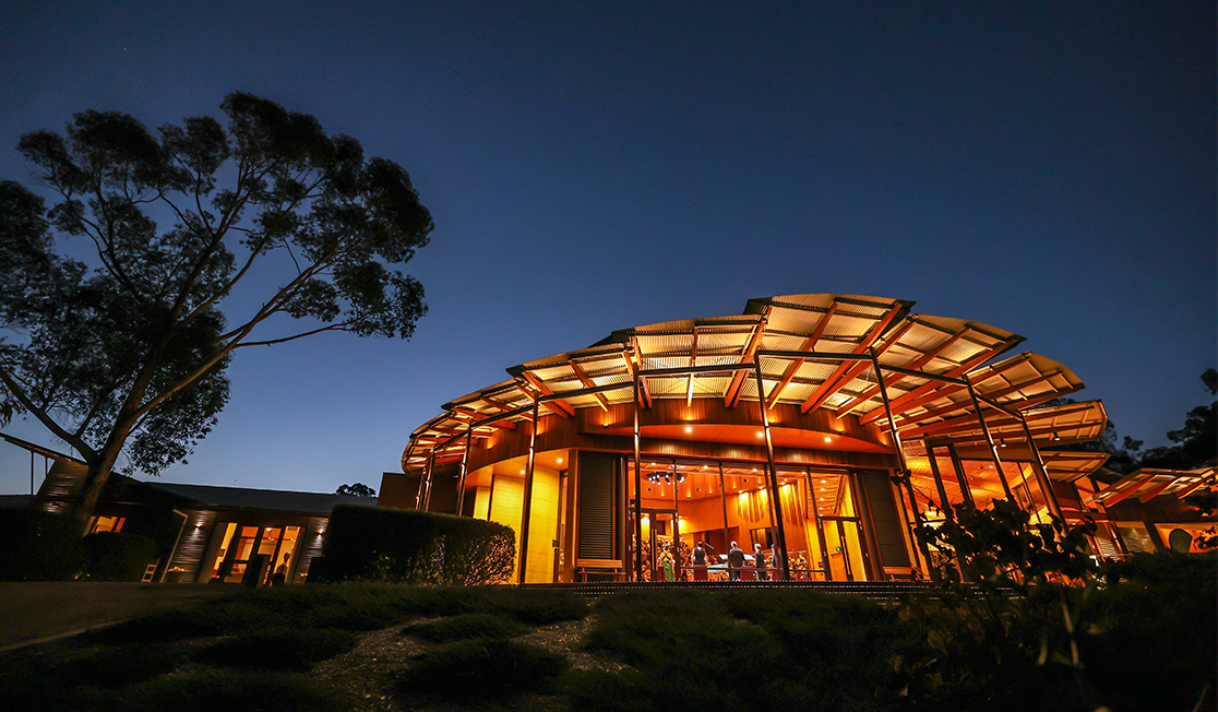 The UKARIA cultural centre building pictured at twilight. There is a large gum tree next to the building. The building is lit by gold light and there is a concert occurring inside the venue and the backs of the musicians are visible.